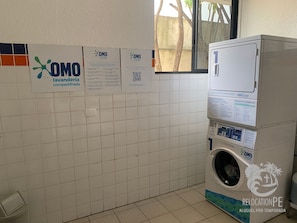 Un espace buanderie équipé d’un lave-linge, agrémenté d’enseignes portant le logo « OMO, laverie partagée », et doté d’un mur carrelé.