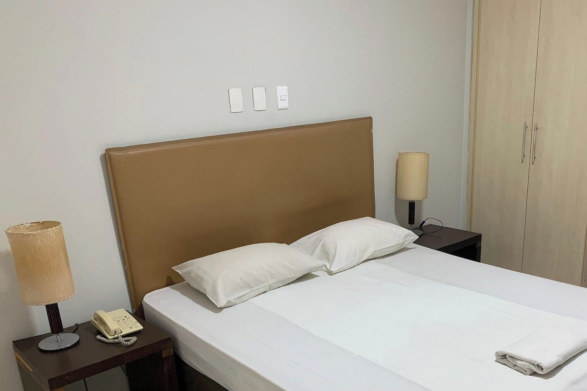 A hotel room with a bed, bedside lamp, phone, and a wardrobe.