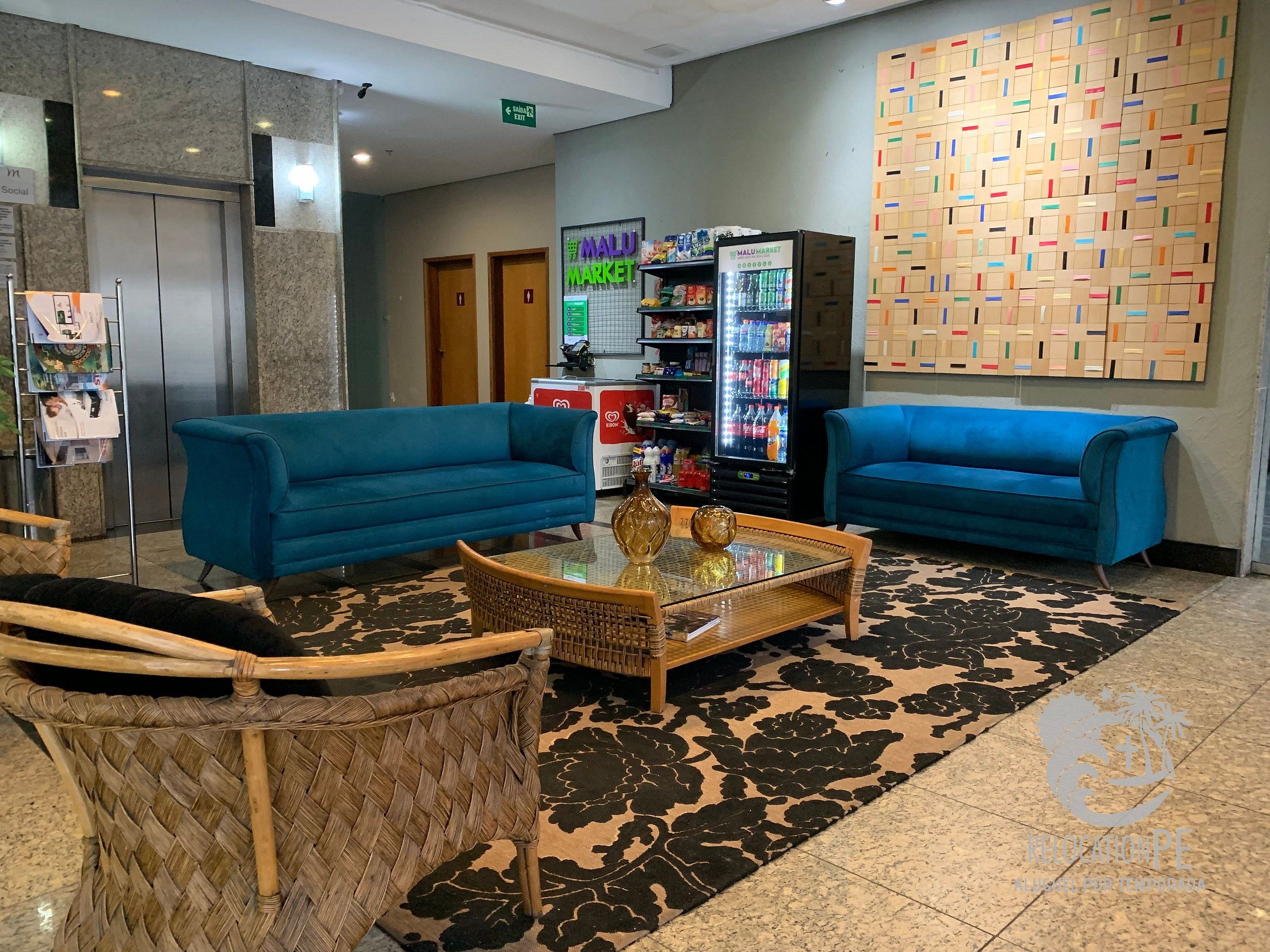 A lobby with two blue sofas, a wicker chair, a glass coffee table, a refrigerator, and a wall with a patterned design.