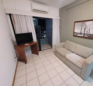 A living room with a television on a wooden stand, a beige sofa, a tiled floor, and an air conditioning unit on the wall.