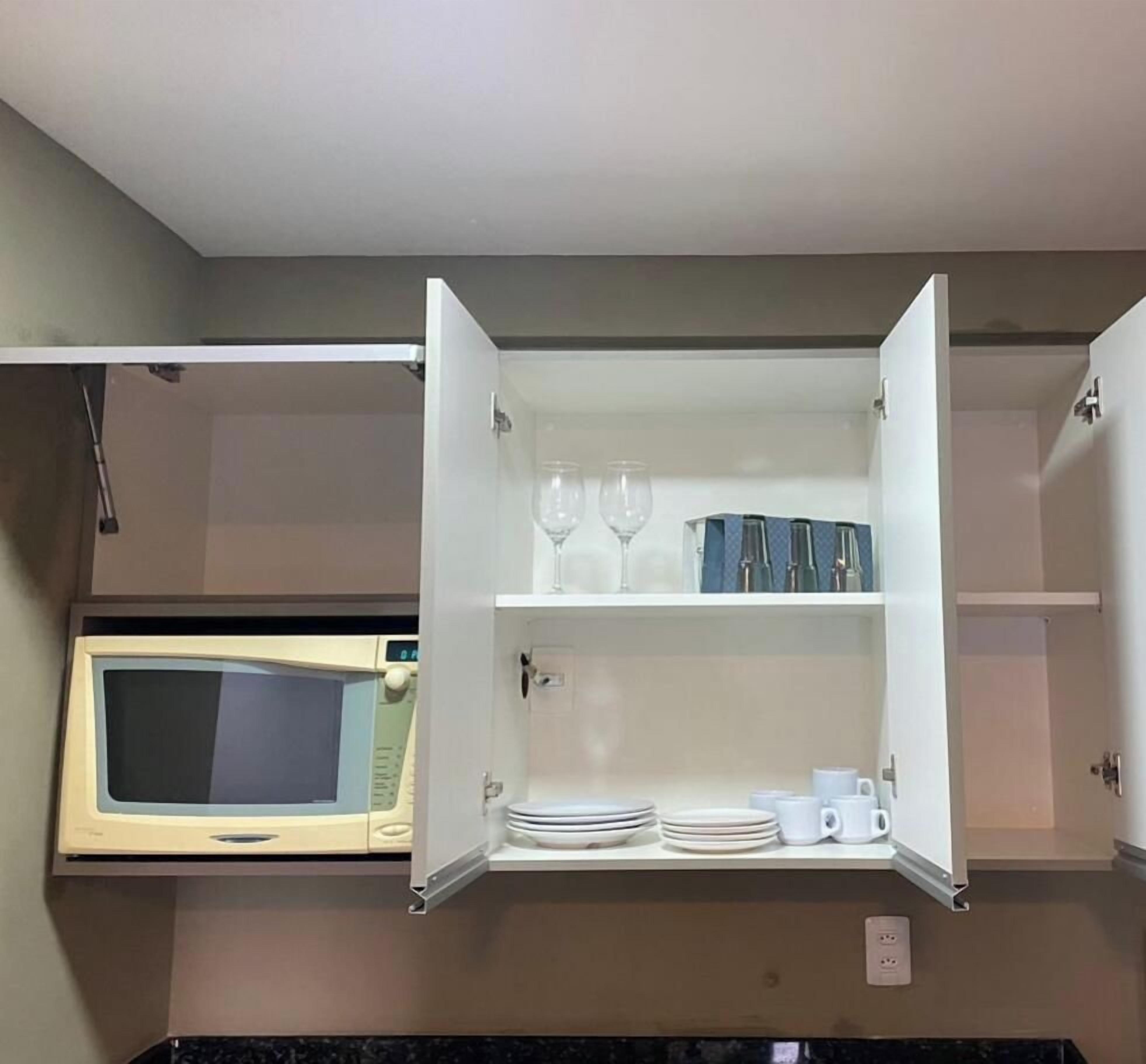 A kitchen with a microwave, a cabinet with dishes and glasses, and a power outlet.