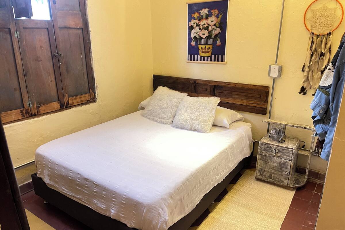 A bedroom with a bed, pillows, a nightstand, a dreamcatcher, and a wall-mounted picture.