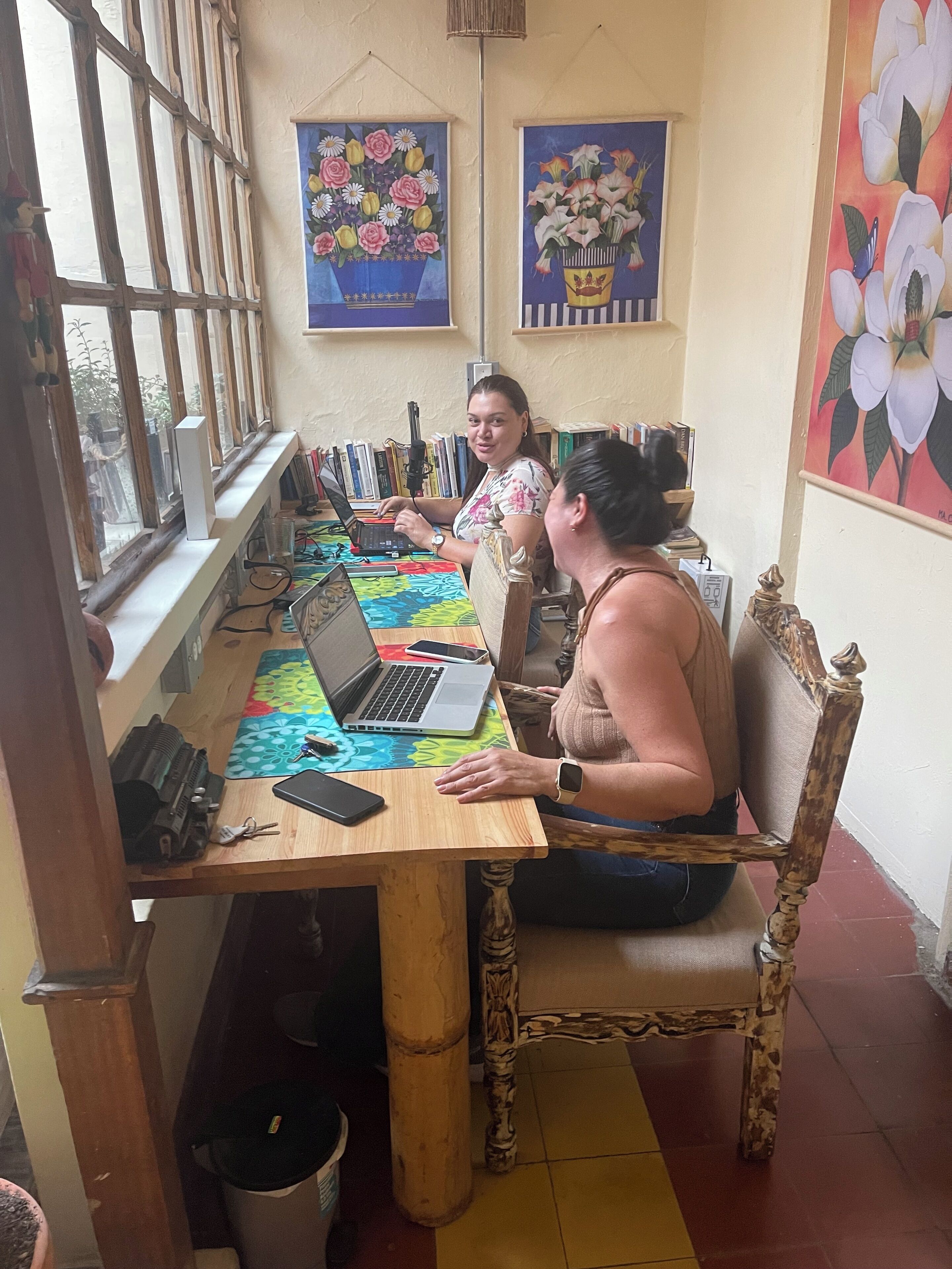 Een kamer met een houten tafel, een laptop en een persoon zittend op een stoel. Er liggen boeken op de tafel en een raam met tralies.