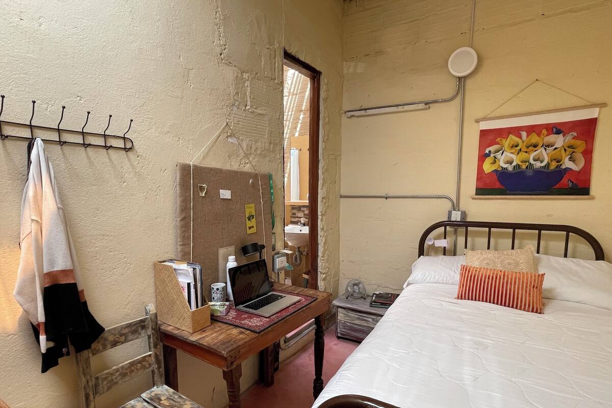 A bedroom with a bed, a wooden desk with a laptop, a chair, a radiator, and a painting on the wall.