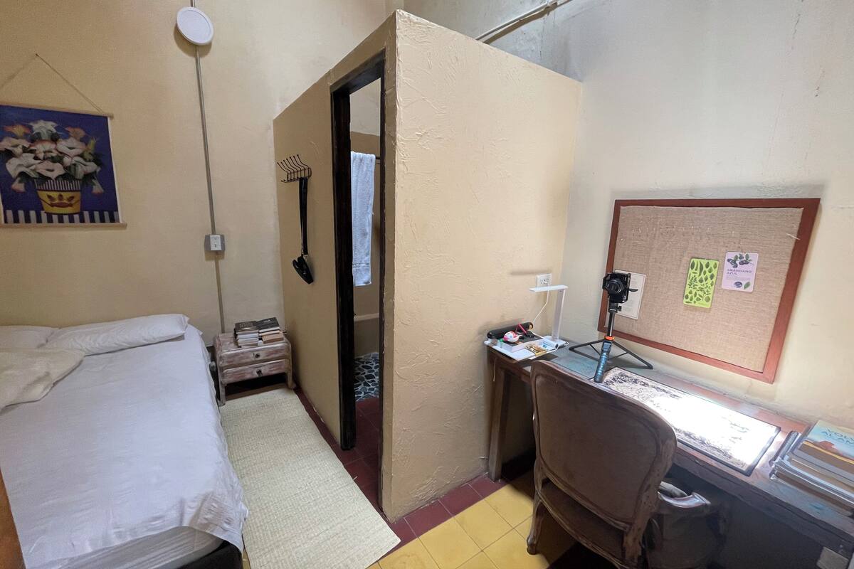 A small, compact hotel room with a single bed, a desk with a computer, a chair, and a corkboard with notes.