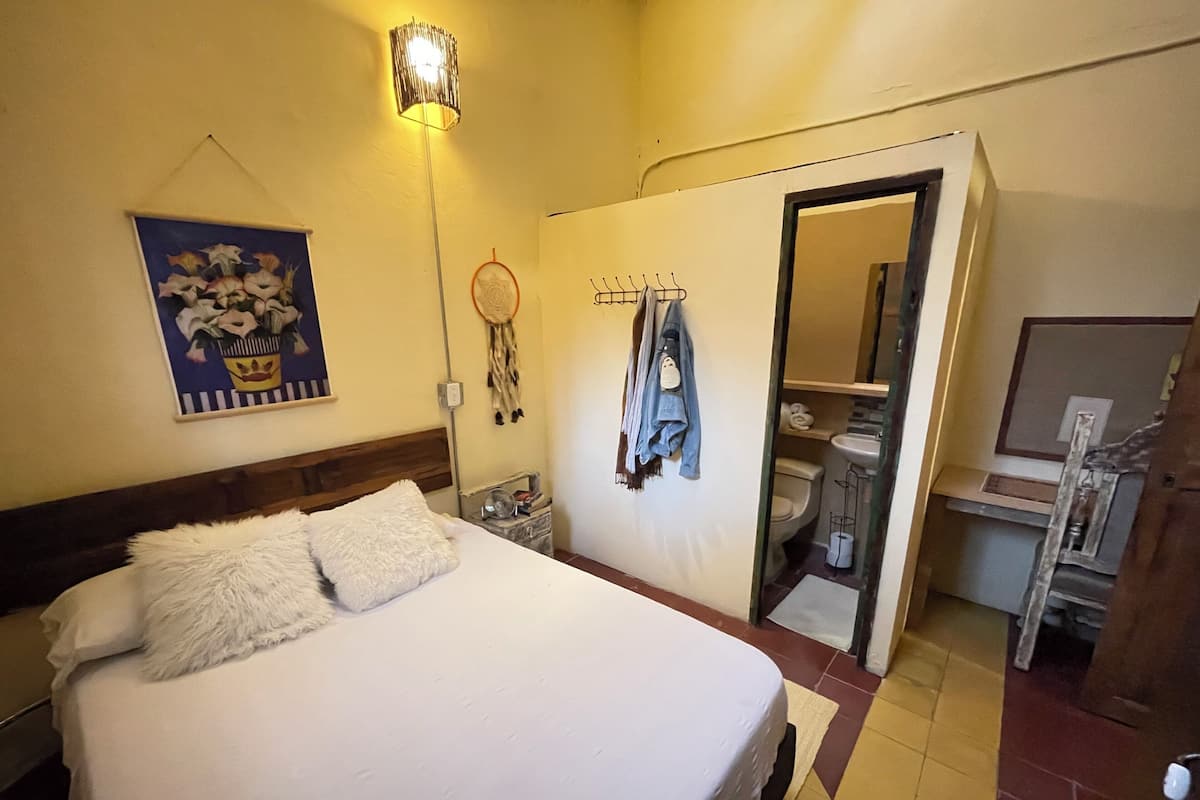 A hotel room with a bed, a nightstand, a wall-mounted lamp, a dreamcatcher, a mirror, and a bathroom visible through an open door.