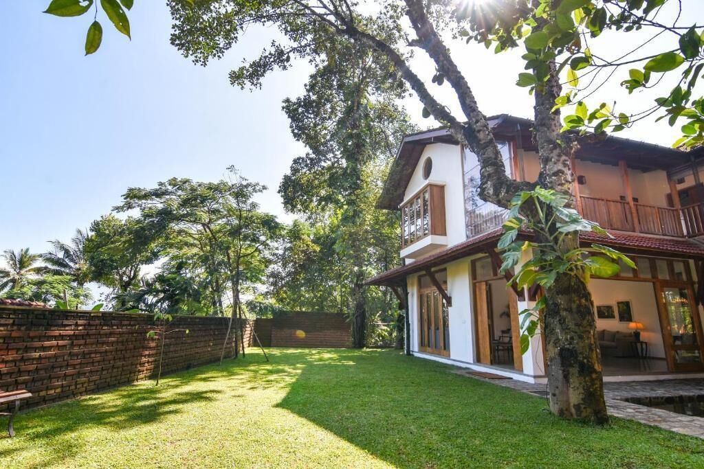 Una casa de dos pisos con balcón, rodeada por un muro de ladrillo y un patio cubierto de césped.