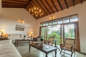 Habitación amplia con una cama grande, un sofá, una mesa de centro y dos sillas. Amplias ventanas ofrecen vistas a zonas verdes.