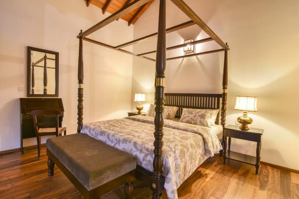 Uma cama com dossel sobre quatro pilares, um banco de madeira, duas mesas de cabeceira com luminárias, uma cadeira e um espelho na parede.