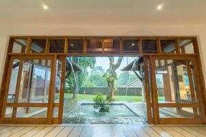 Puerta de cristal con marco de madera que da a un jardín con una pequeña fuente y un árbol.