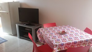 Ein kleines Zimmer mit einem Fernseher auf einem Schrank, einem Tisch mit einer bunten Tischdecke und roten Stühlen.