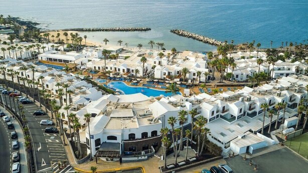 Vista aérea de un complejo turístico costero con varios edificios blancos, una piscina central y vista al mar.