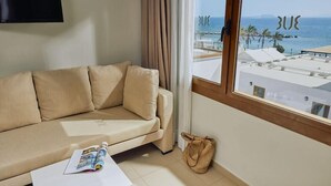 Un sofá color beige con una revista, un televisor montado en la pared y vista al mar a través de una ventana.