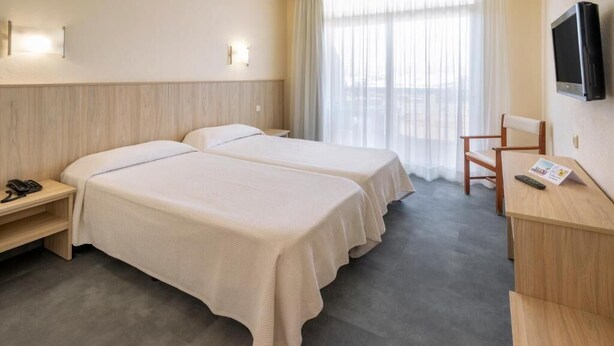 A hotel room with two beds, a desk, a chair, a TV, and a window with curtains. - Marinada Hotel (Salou)
