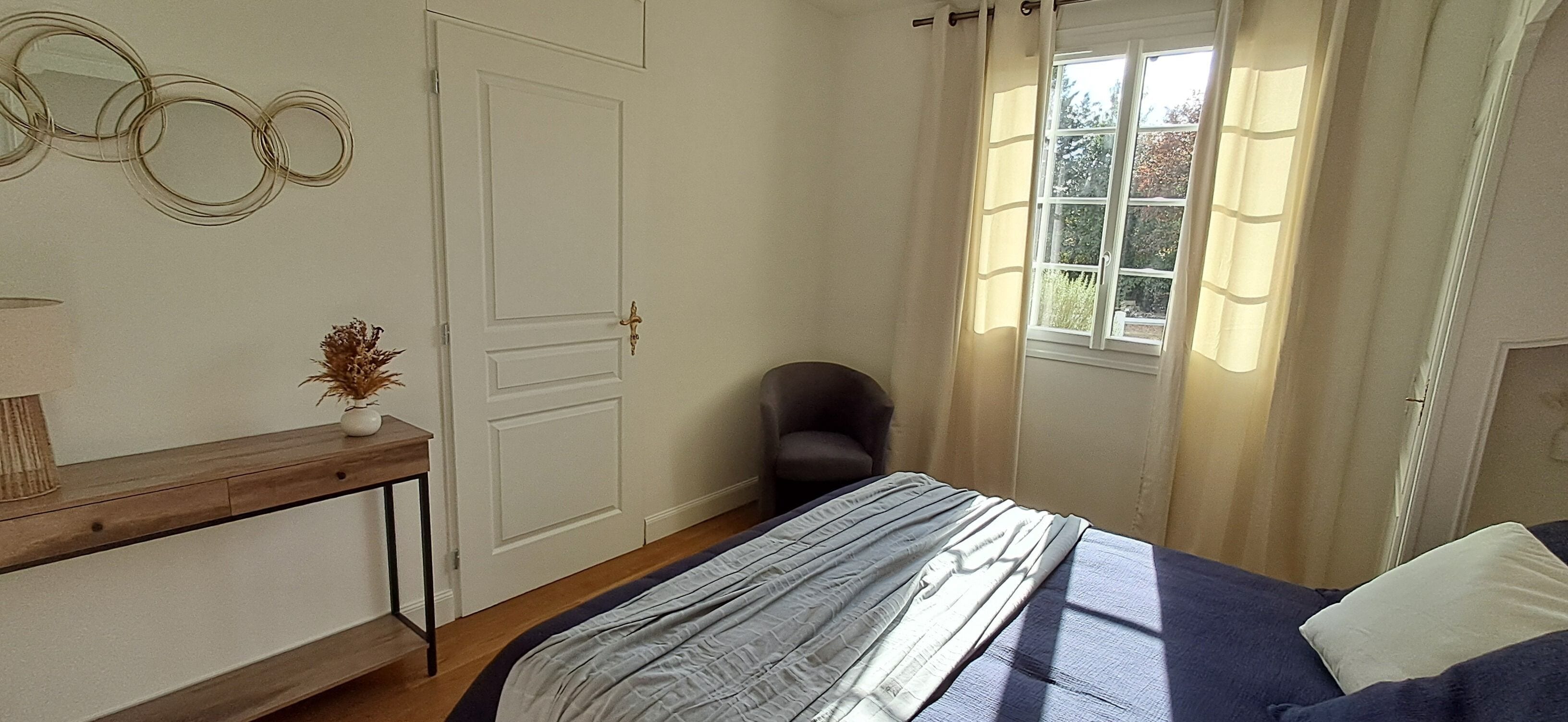 A bedroom with a bed, a bedside table with a lamp, a chair, a window with curtains, and a door.