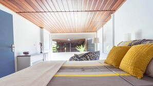 A modern bedroom with a wooden ceiling, a bed with patterned bedding, and a view of a balcony with plants. - Very nice spacious apartment with Jacuzzi - Le Canopé (Le Gosier)