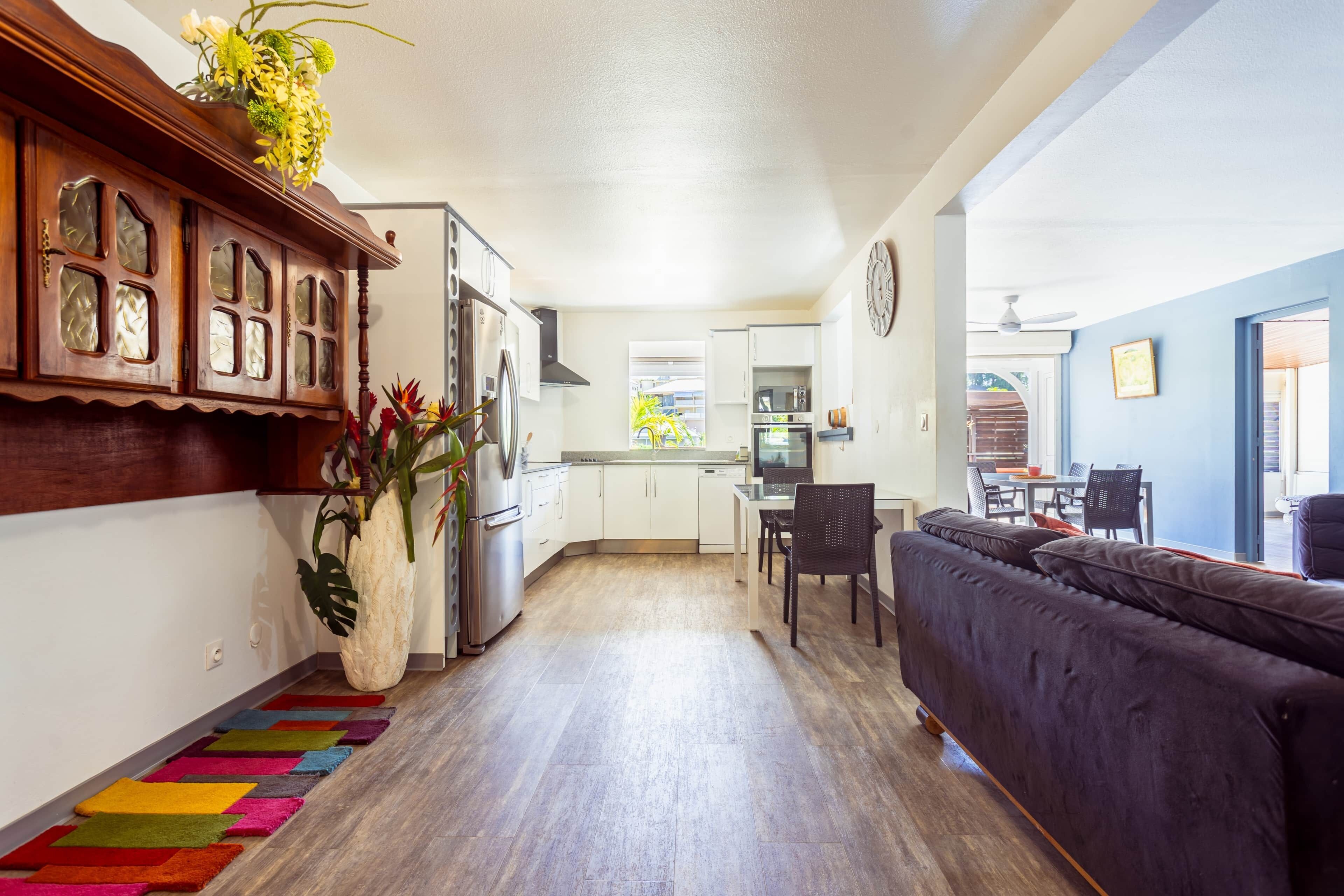 A modern kitchen with white cabinets, a dining area with a dark sofa, and a colorful rug.