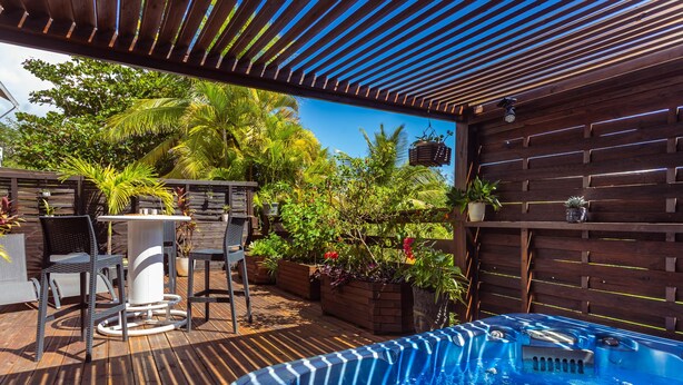 A wooden deck with a hot tub, outdoor furniture, and lush greenery. - Very nice spacious apartment with Jacuzzi - Le Canopé (Le Gosier)