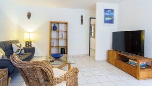 Ein Wohnzimmer mit einer Couch, einem Glastisch, einem Korbstuhl, einer TV-Konsole mit Flachbildfernseher, einem Bücherregal aus Holz und einer Wandhalterung.
