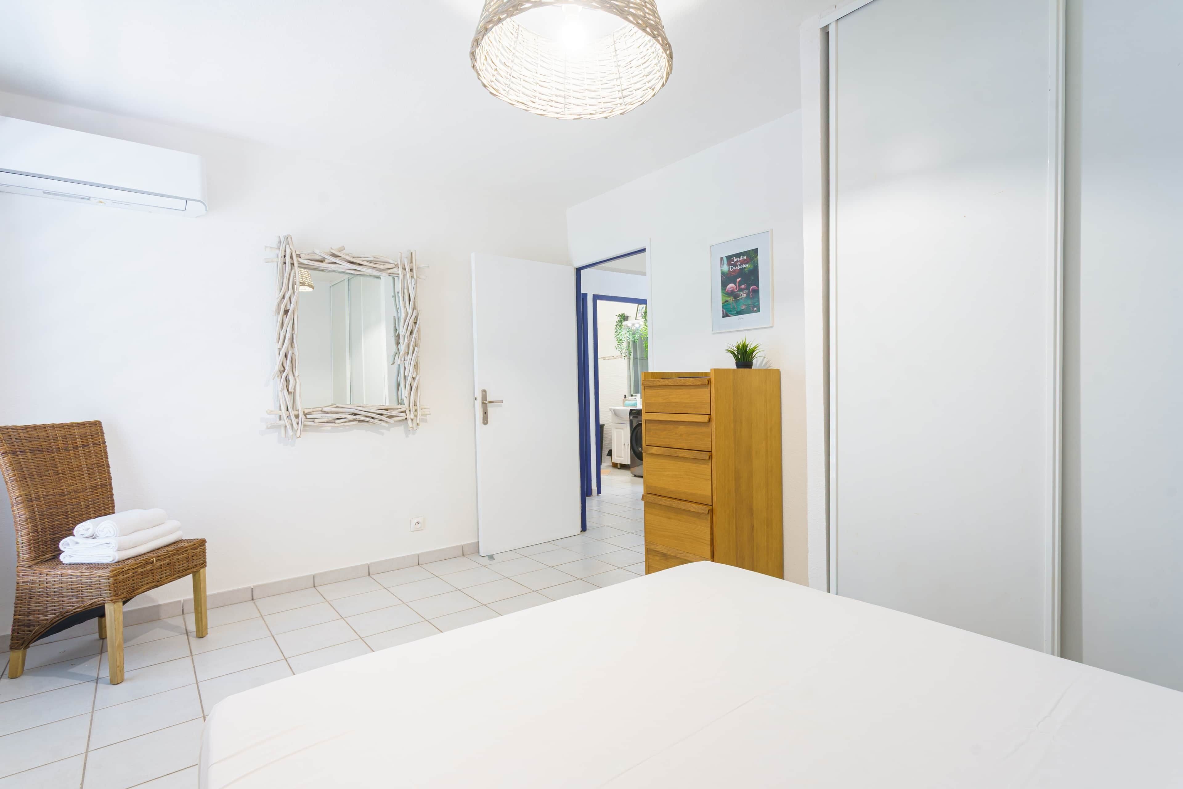 A modern hotel room with a large bed, a wooden dresser, a wicker chair with towels, a mirror, and a door leading to another room.