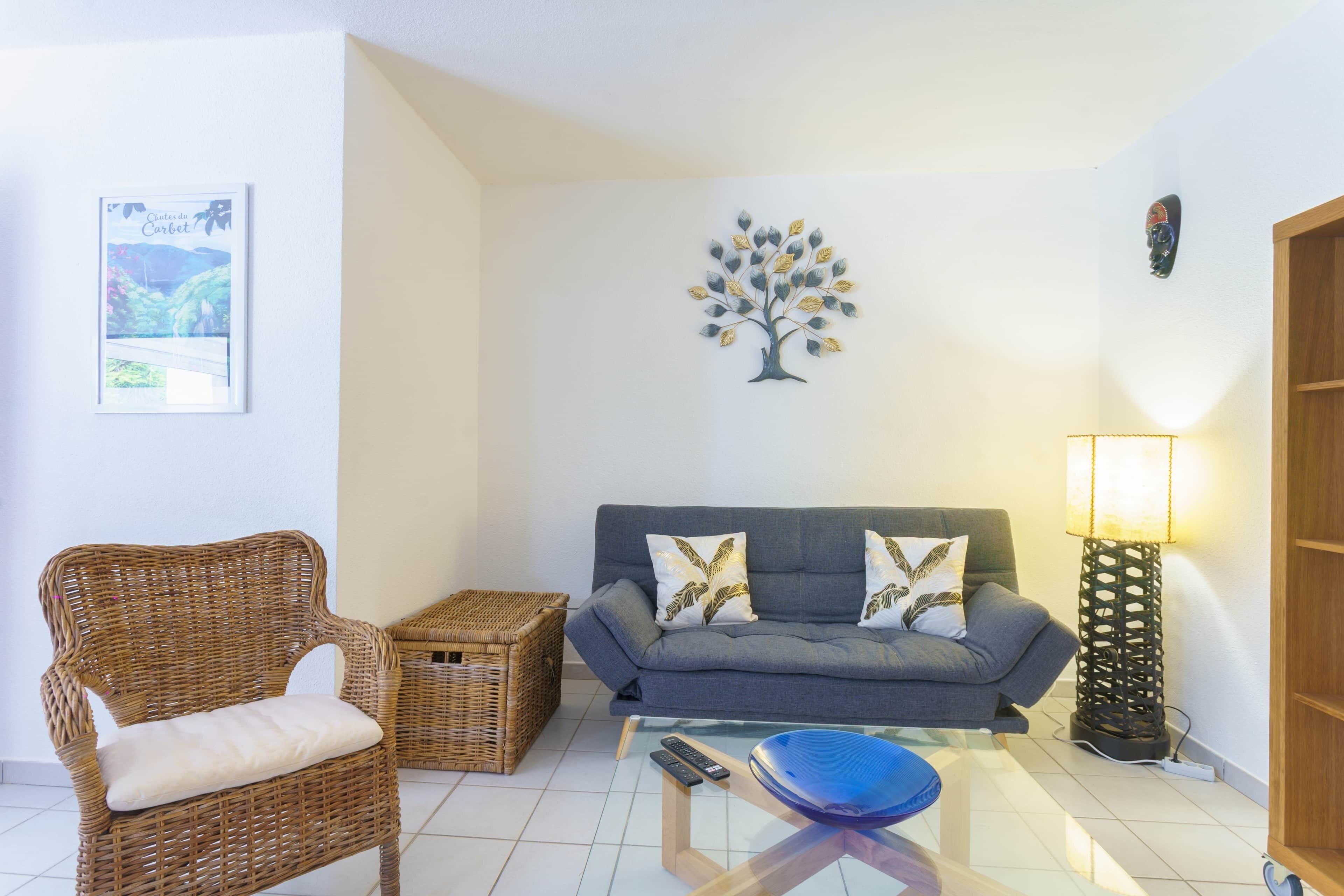 Ein Wohnzimmer mit einem Korbstuhl, einem grauen Sofa mit Zierkissen, einem Glastisch mit blauer Schale, einem gerahmten Bild an der Wand und einer Stehlampe.