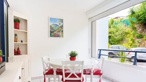 Ein Essbereich mit einem runden Tisch, weißen Stühlen und einem roten Kissen. Es gibt eine weiße Schrank mit Regalen, eine Topfpflanze und ein gerahmtes Bild an der Wand.
