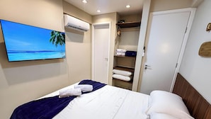 Ett hotellrum med en säng, en tv som visar en strandscen, en luftkonditioneringsanläggning och en garderob med hopvikta handdukar.