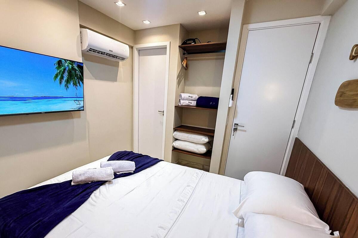 Une chambre d’hôtel avec un lit, un téléviseur diffusant une scène de plage, un appareil de climatisation et une armoire contenant des serviettes pliées.