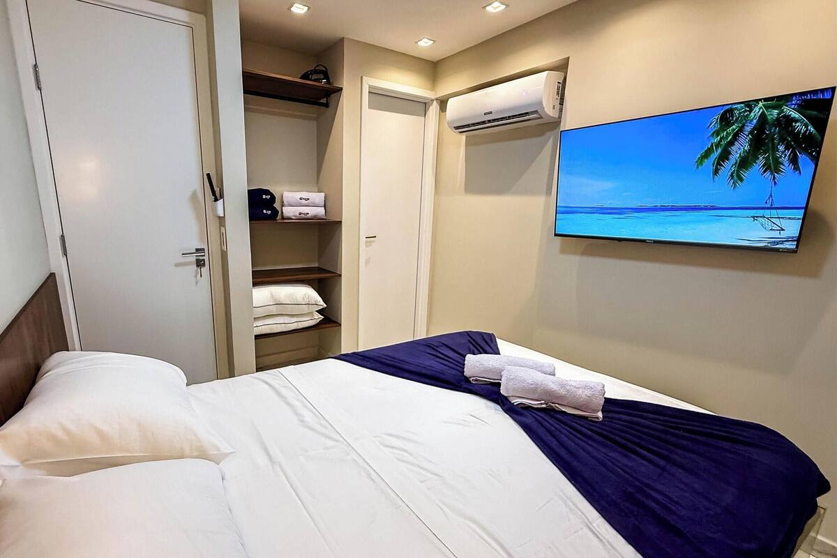 Une chambre d’hôtel équipée d’un lit, d’une armoire, d’un appareil de climatisation et d’un téléviseur diffusant des images d’une plage.