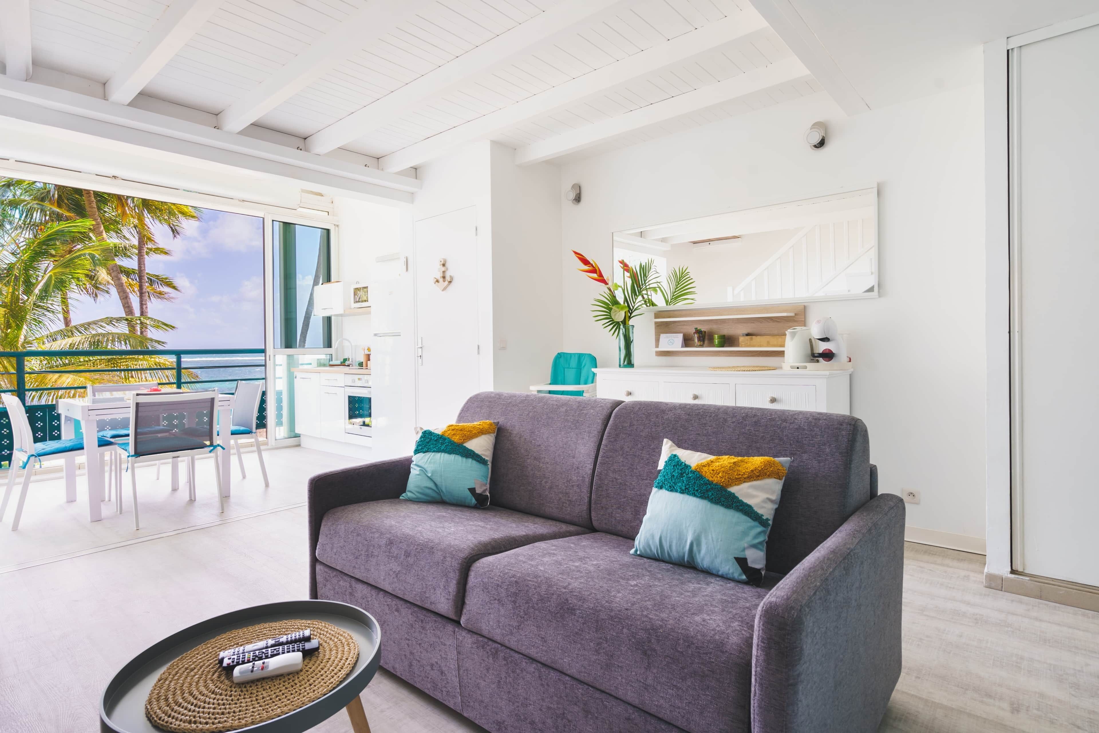 Una sala de estar moderna con un sofá gris, un comedor con sillas blancas, una cocina y un ventanal con vistas a palmeras.