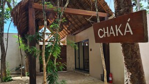 Une enseigne en bois portant le nom « CHAKA », dans un décor rustique avec une structure à toit de chaume, un sol en pierre et une végétation luxuriante tout autour.