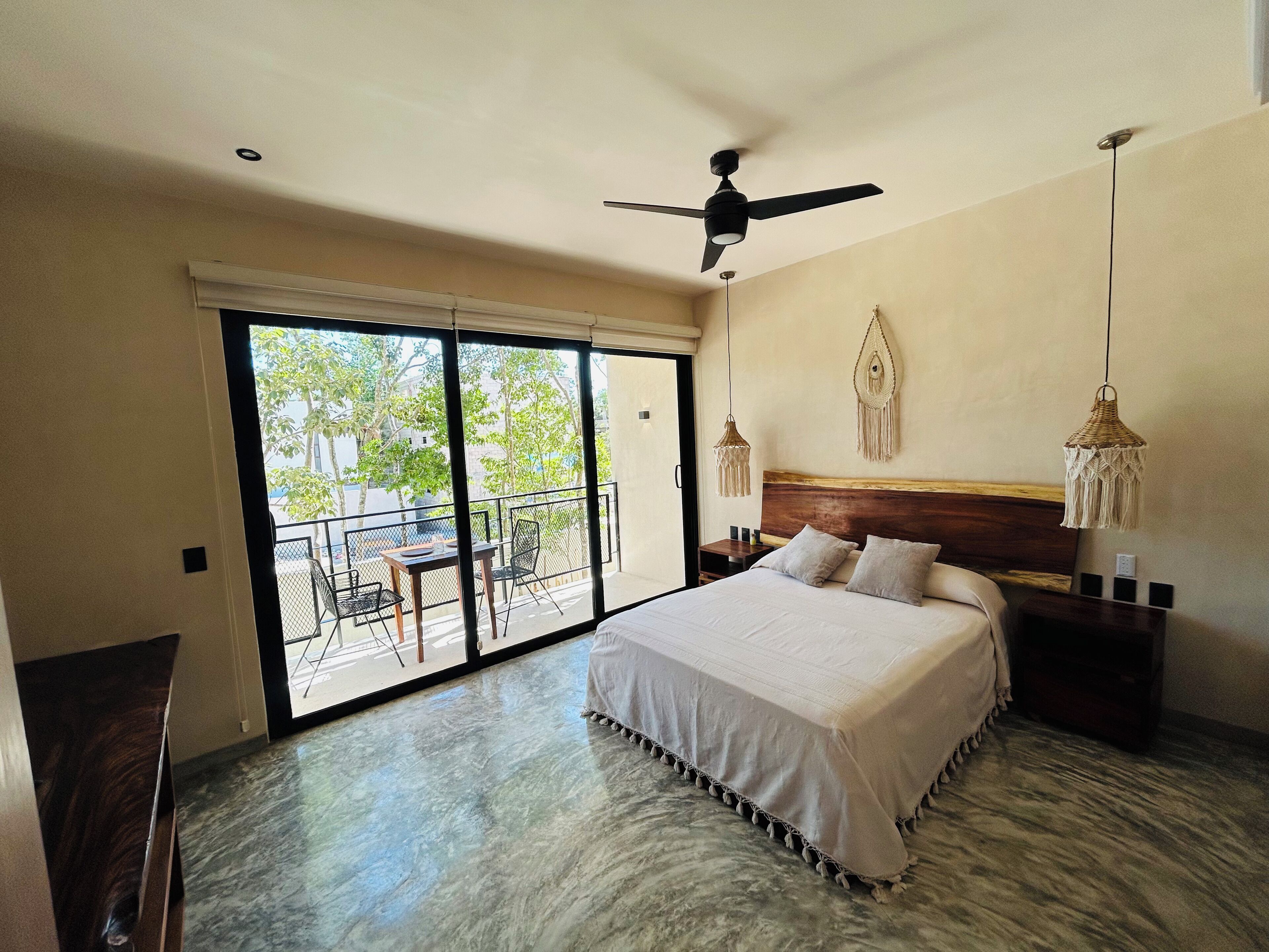 A bedroom with a large bed, a ceiling fan, a balcony with a table and chairs, and a macrame wall hanging.