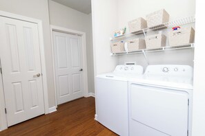 A laundry room with a front-loading washer and dryer, white doors, and storage shelves with baskets. - Rebel Bluff on Depot by Apex Short Term Stays (Oxford)