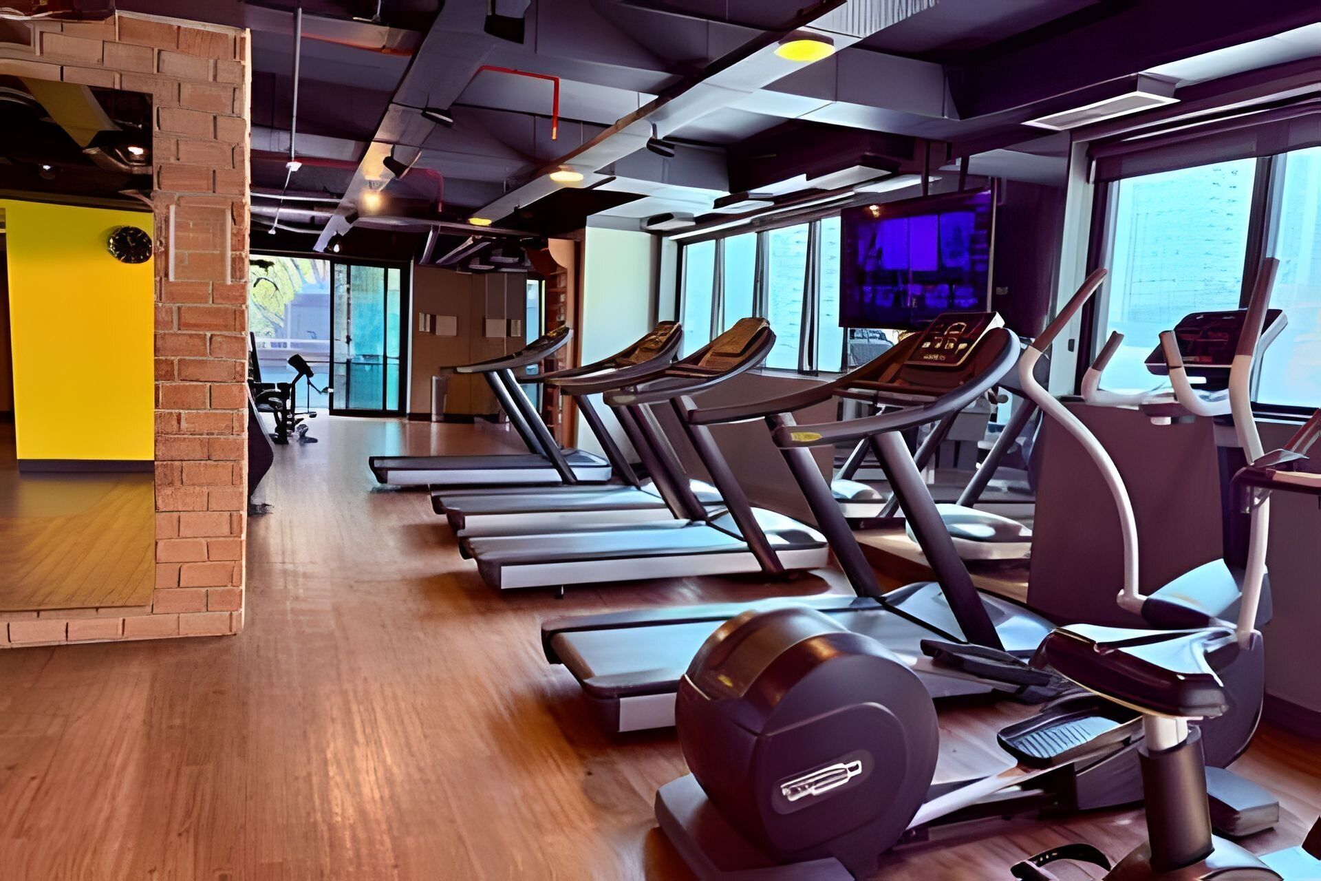 A gym with multiple treadmills and elliptical machines, wooden flooring, and large windows offering views of the outside.