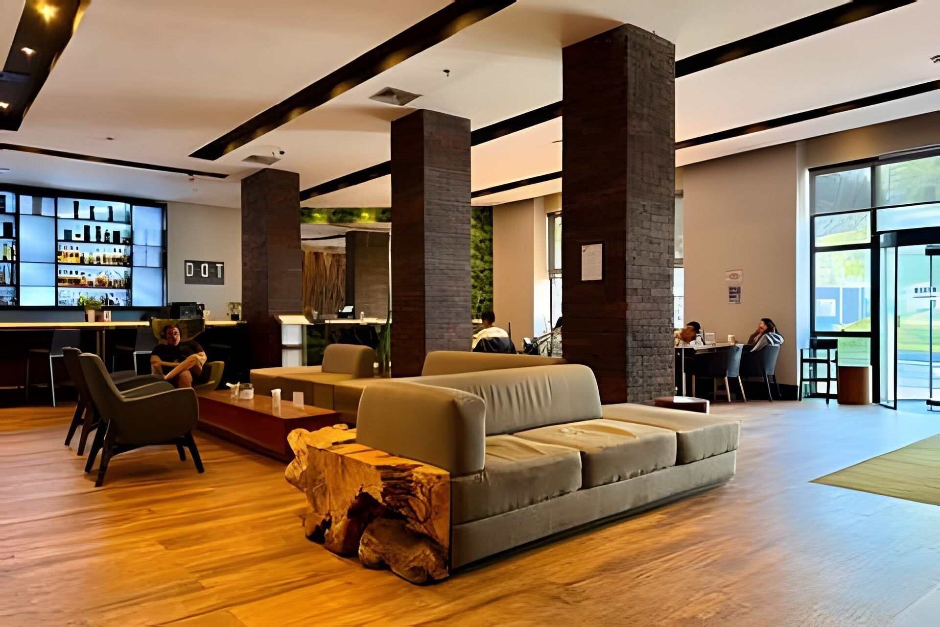 A modern lobby with a wooden floor, a grey sofa, a bar area with shelves, and a large window providing a view of the outdoors.