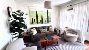 A living room with a sofa, armchairs, a coffee table, a large painting, a potted plant, and a clock on the wall. - Casa Italiana: Cozy home in the heart of The Hill (St. Louis)