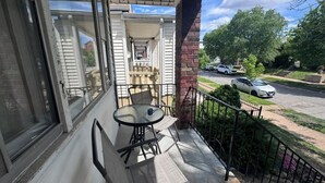 A balcony with a table and chairs, a view of a street with parked cars, and a house in the background. - Casa Italiana: Cozy home in the heart of The Hill (St. Louis)