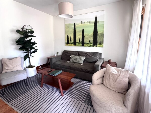 A living room with a grey sofa, a coffee table, a striped rug, a large potted plant, and a painting of trees. - Casa Italiana: Cozy home in the heart of The Hill (St. Louis)