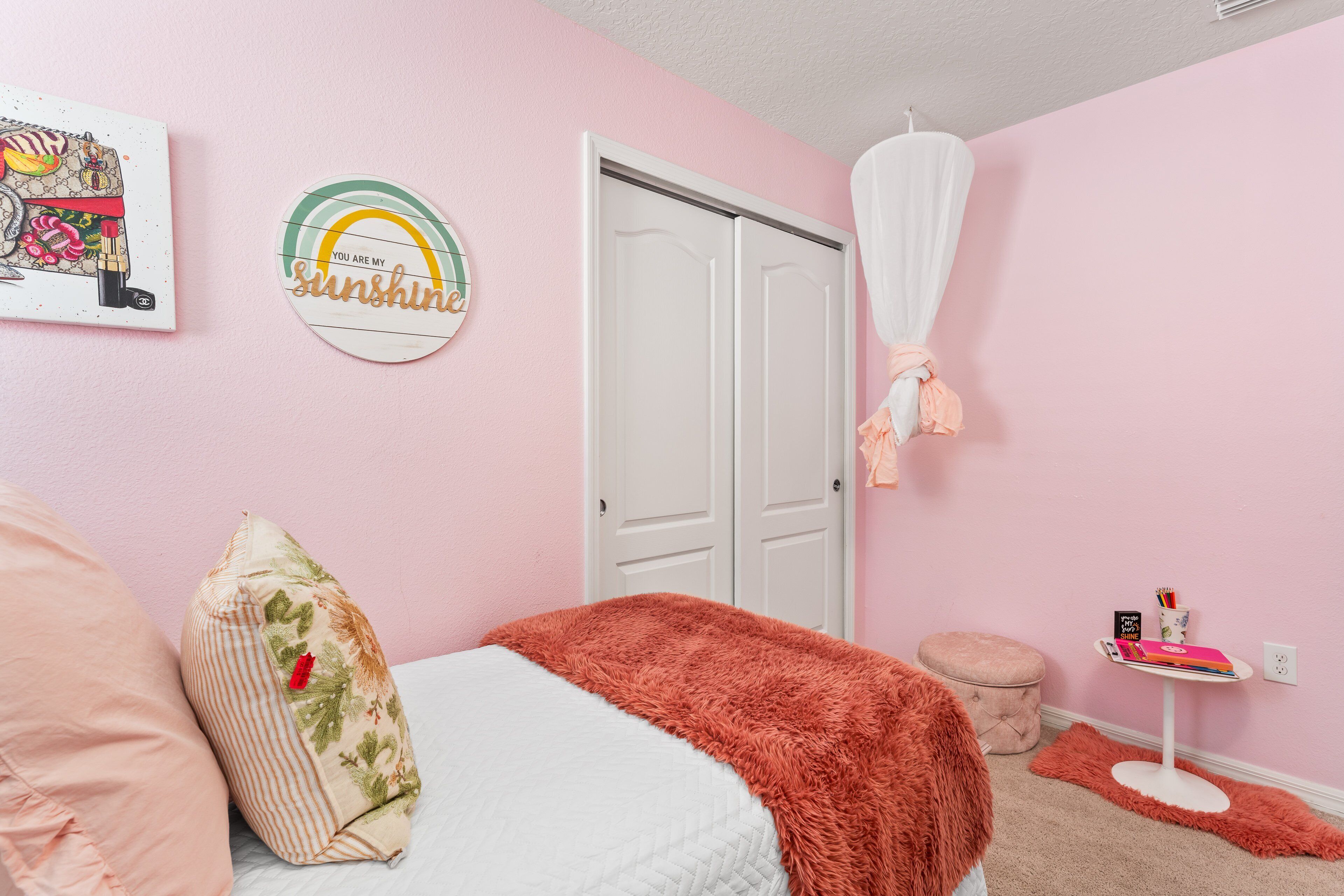 A pink-themed bedroom with a bed, pillows, a closet, and a small table with books and a cup.