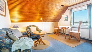 A cozy room with a wooden ceiling, a sofa, a chair, a small table, and a window with curtains. - Apartment Barlach, 35 sqm, 1 Bedroom, Max. 2 Guests (Pellworm)