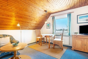 A room with a sloped wooden ceiling, a dining table with chairs, a sofa, a television, and a window with curtains. - Apartment Barlach, 35 sqm, 1 Bedroom, Max. 2 Guests (Pellworm)