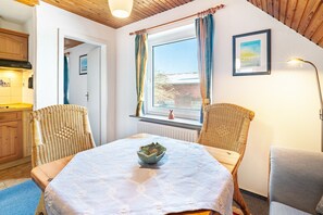 Une pièce chaleureuse avec une table à manger, deux chaises en osier, une fenêtre avec des rideaux, un tableau au mur et une lampe.