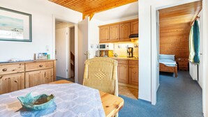 A compact room with a wooden ceiling, a dining table with a bowl, a wicker chair, a bed, and a kitchenette with wooden cabinets. - Apartment Nolde, 33 sqm, 1 Bedroom, Max. 2 Guests (Pellworm)