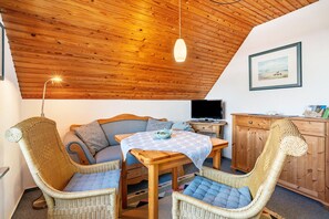 Une pièce chaleureuse avec un plafond en bois, des meubles en osier, une table recouverte d’une nappe bleue et un téléviseur.
