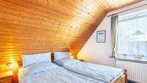 A room with a sloped wooden ceiling, a bed with white bedding, a bedside table, and a window with a view of a snowy landscape. - Apartment Nolde, 33 sqm, 1 Bedroom, Max. 2 Guests (Pellworm)