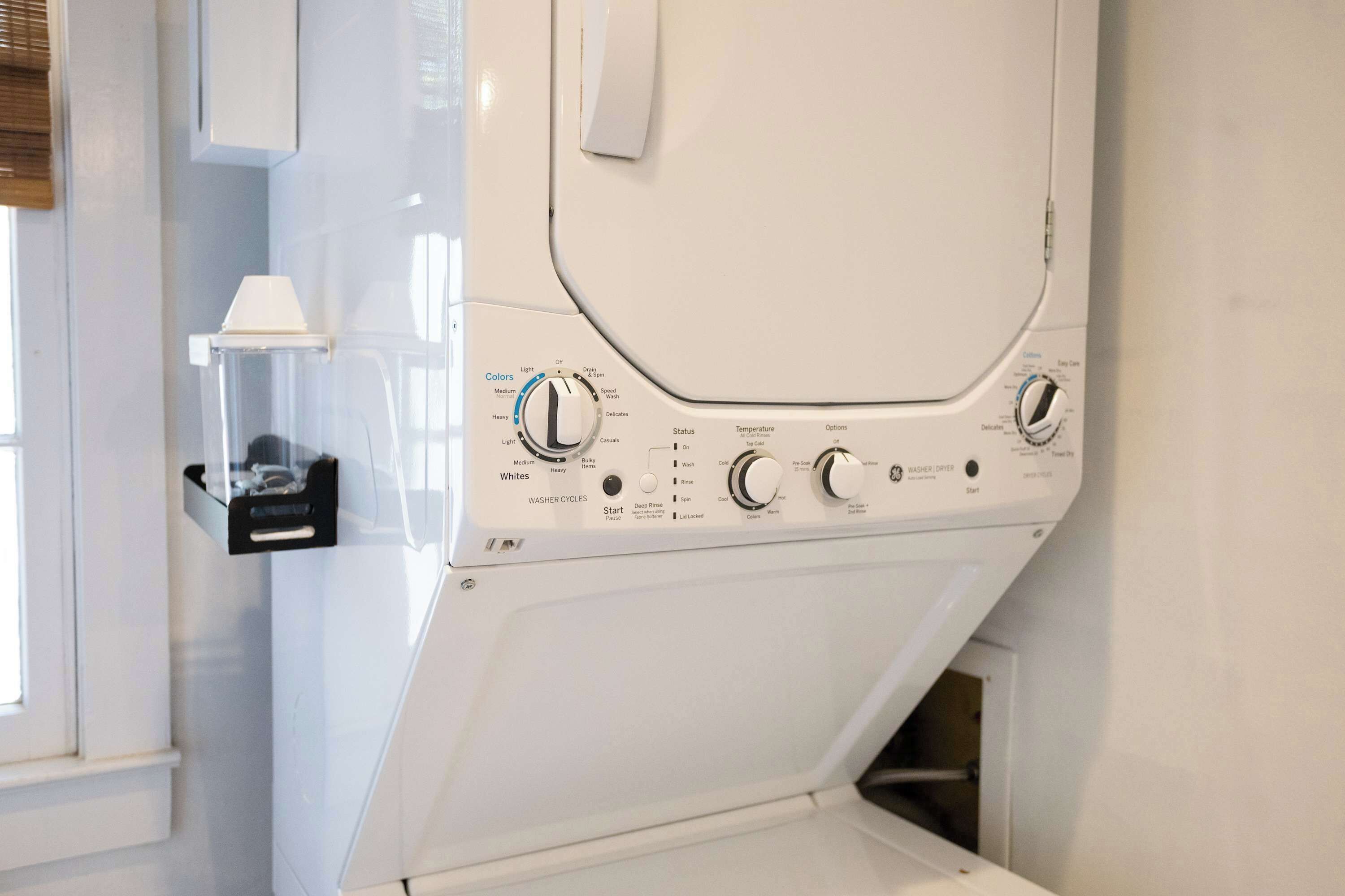 A front-loading washing machine with control knobs, a detergent drawer, and a small shelf with a white container.