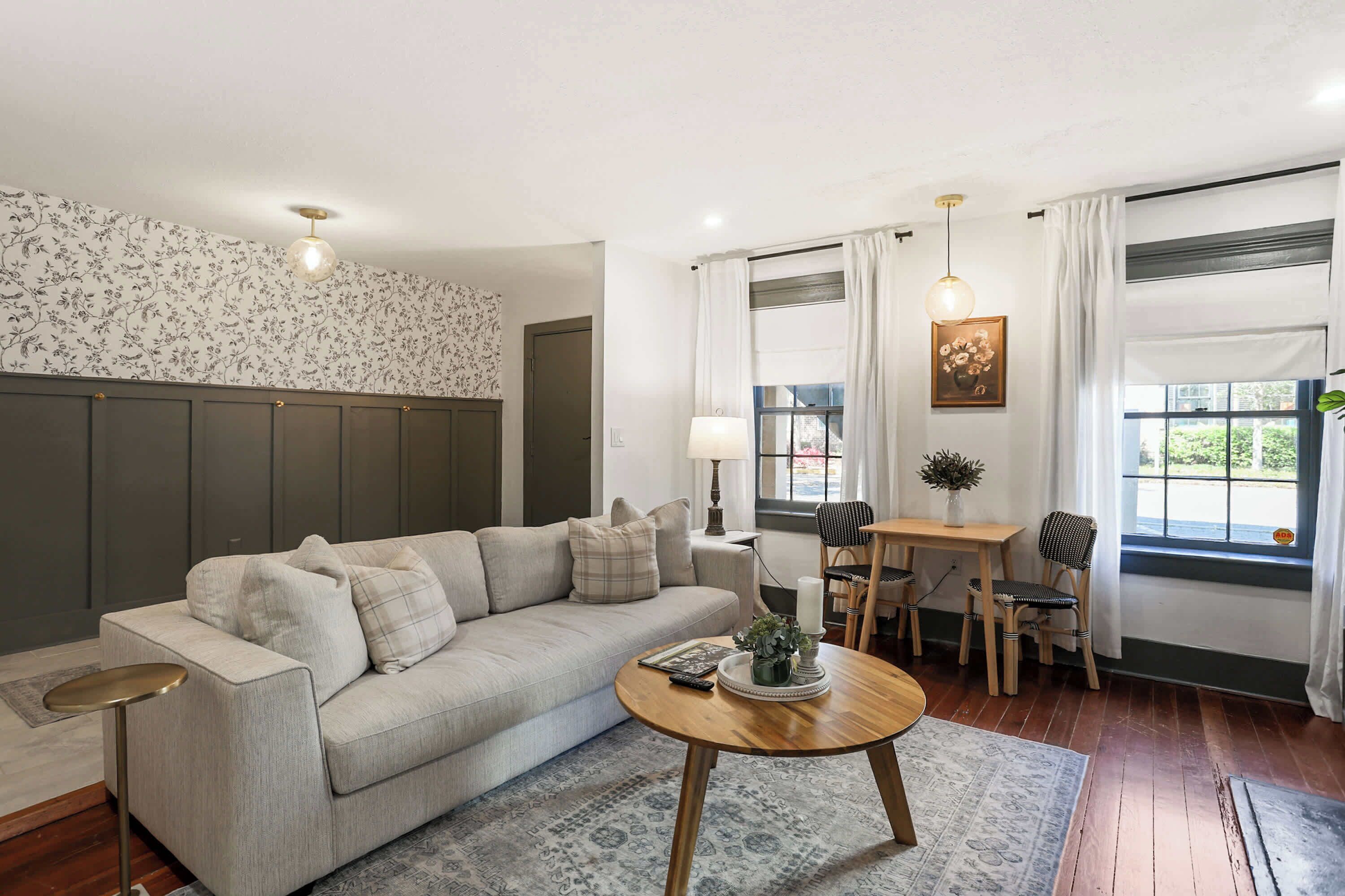 Een woonkamer met een bank, een rond salontafeltje, een eettafel met stoelen, een lamp en een raam met gordijnen.