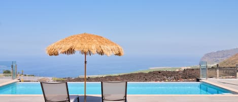 A pool area with two lounge chairs and a thatched umbrella, overlooking a coastal landscape.