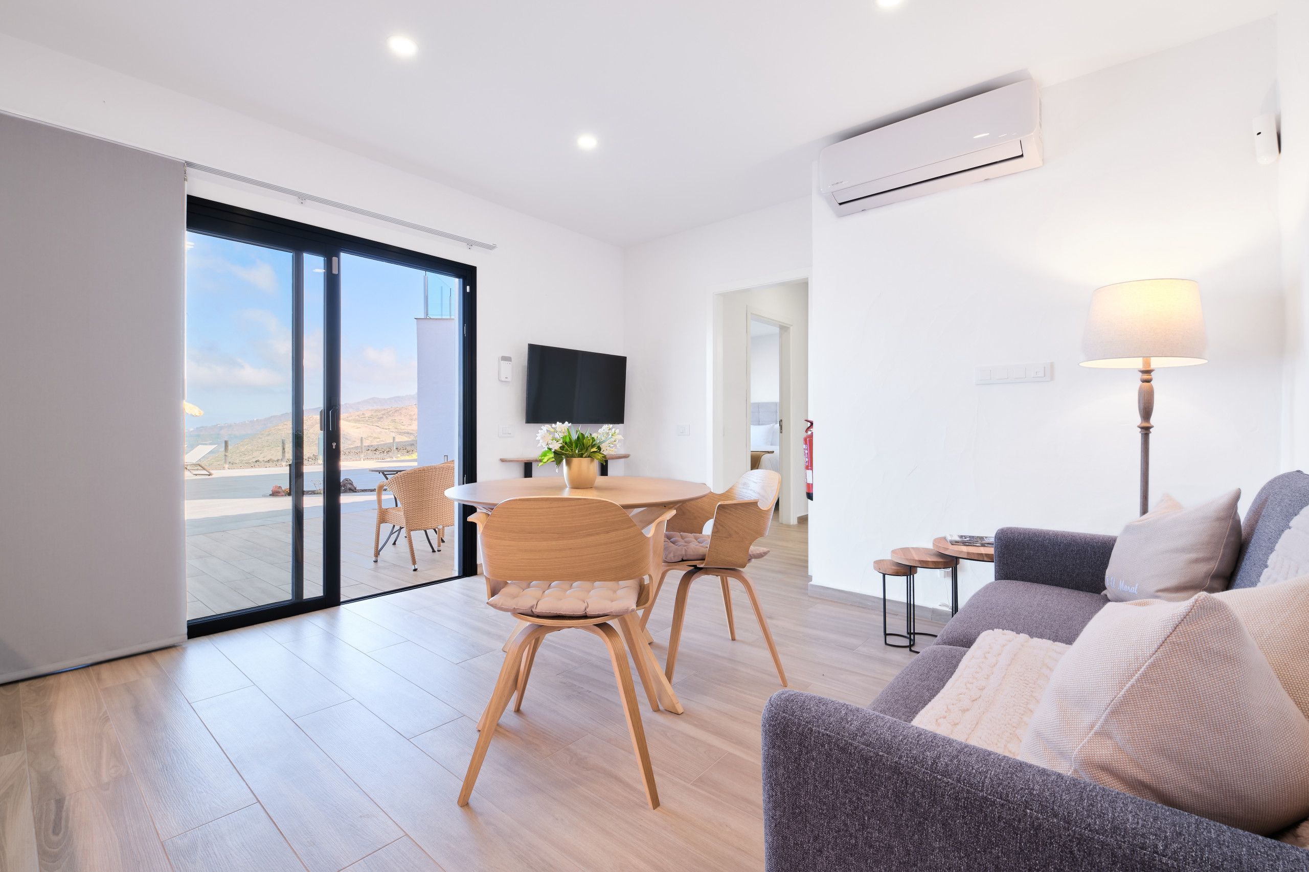 A modern living room with a sofa, a dining table with chairs, a TV mounted on the wall, and a large sliding glass door leading to an outdoor area.