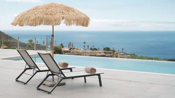 Un espace aménagé au bord de la piscine, avec deux chaises longues et un parasol en paille, offrant une vue sur une mer d’un bleu limpide.
